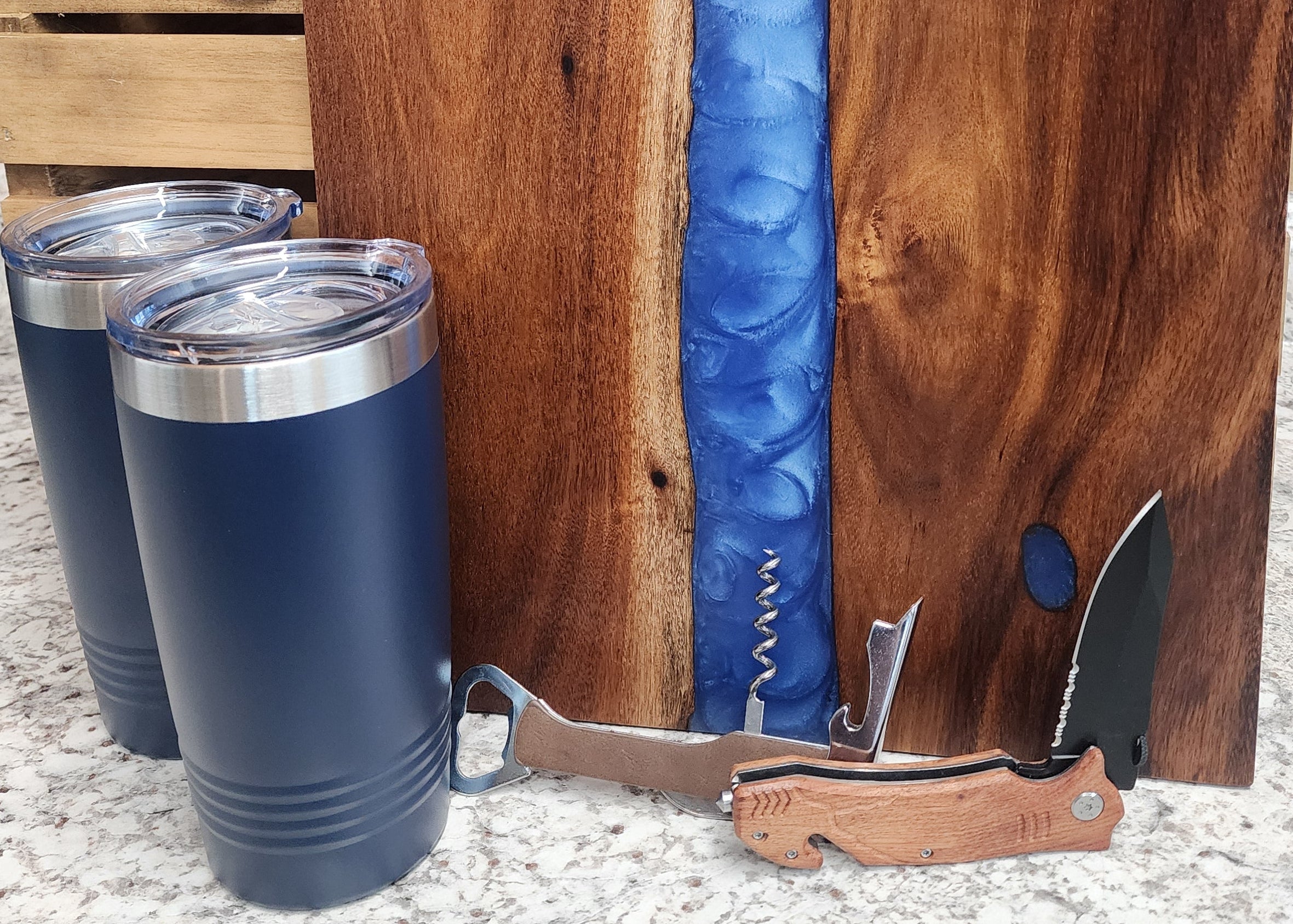 Wooden cutting board with blue resin inlay and a knife, next to a blue tumbler on a textured surface.